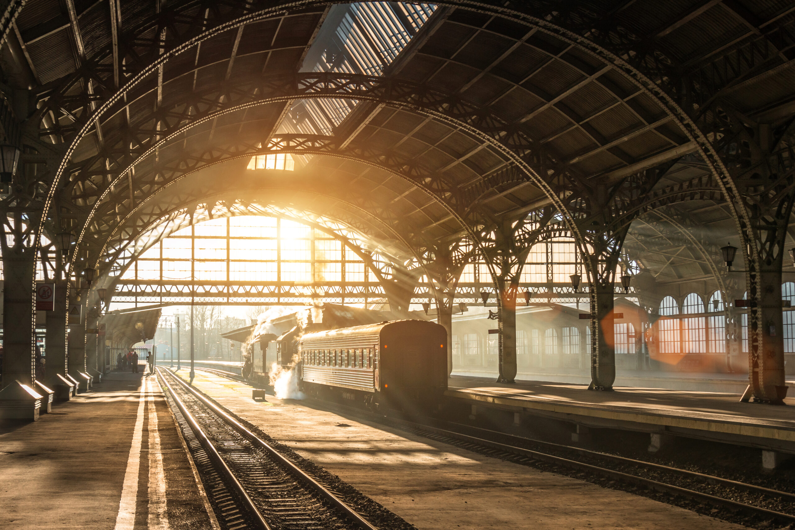 Old railway station with a train and a locomotive on the platform awaiting departure. Evening sunshine rays in smoke arches.